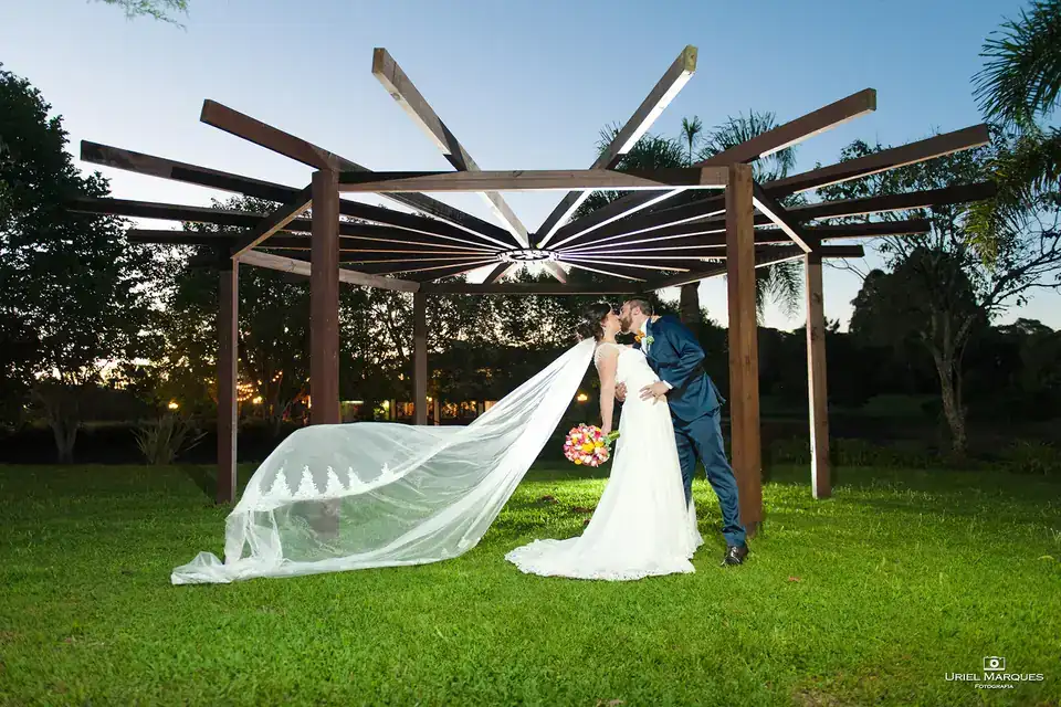 Noivos se beijando sob pérgola de madeira, véu esvoaçante ao entardecer